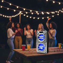 Silver double subwoofer bluetooth speaker with blue LED glow at an outdoor backyard party at night